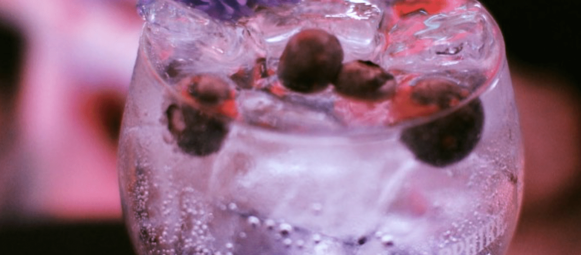 A vibrant purple gin and tonic cocktail served in a tall glass with ice cubes and fresh blueberries, garnished with a slice of lime and herbs.